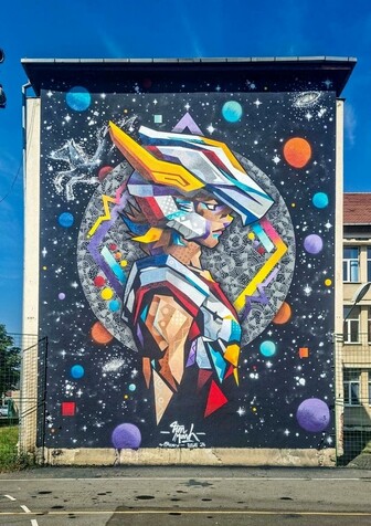 Streetartwall. A large mural of a manga character with armor and a bird helmet was sprayed/painted on the outside wall of a three-story modern building. It is Seiya, a warrior of sorts, depicted in colorful armor composed like a mosaic of colors and patterns. He looks to the side and is surrounded by colorful planets, moons and a starry sky. The constellation of Pegasus can be seen above him.
Info: Saint Seiya (Japanese: 聖闘士星矢, Seinto Seiya) is a globally successful manga series by Japanese illustrator Masami Kurumada. The characters are reincarnations of Greek gods and mythical figures. The given theme of the street art festival was "Connection between generations", and this wall used to feature an old painting of a flying Pegasus.
