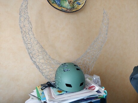 The beginnings of a replica of the False Knight helmet from the game Hollow Knight. A green bike helmet is laying on a stack of magazines and papers. Two large horns made from chicken wire is attached on each side of the helmet and curve upwards in a large arch.
