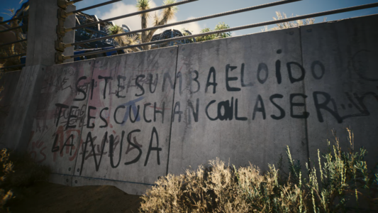 Una pared de cemento gris, parcialmente cubierta por grafitis en español, se encuentra al aire libre bajo un cielo parcialmente soleado.  Los grafitis son ilegibles en su mayor parte, pero se pueden distinguir frases como "SI TE SUNBA EL OIDO TE ESCUCHAN CONLASER LA NUSA". En la parte superior de la imagen, se puede ver una cerca de metal y algo de vegetación desértica baja.