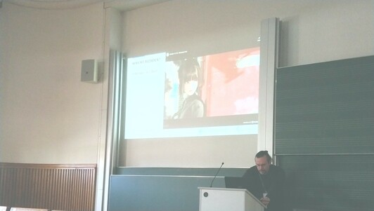 Eine 3D Szene ist in der Präsentation an der Wand zu sehen, der Vortragende schaut von seinem Pult dorthin. Wir sind in einem Hörsaal