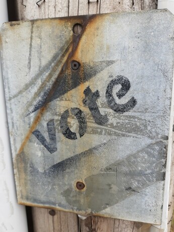 Photo of sheet metal bolted to a street post. The original sign is gone, instead someone has stencil-sprayed VOTE in black paint on the raw metal rectangle.