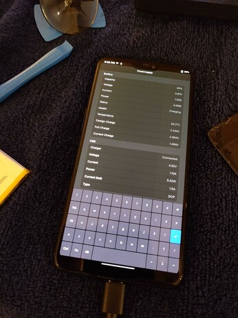 The phone, now powered up and booted into postmarketOS Phosh, with the Powersupply app open showing the battery charging at around 5W and the reported full capacity showing 2.96Ah.