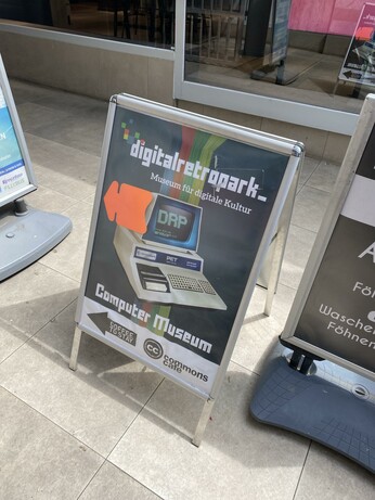A sandwich board sign for "digitalretropak-," a museum celebrating digital culture, specifically a Computer Museum. It features an image of a vintage computer and includes the text in German, indicating it's a place for retro digital exhibitions and possibly a café