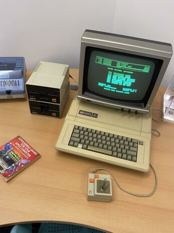 An old computer setup featuring a vintage monitor displaying green text, a beige keyboard, and a joystick. Two external disk drives are visible next to the monitor, along with a book titled "Astounding Games for Your Apple Computer." The scene is set on
