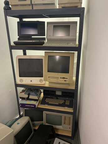 A shelving unit displaying various vintage computers and monitors, including several Apple Macintosh models. The items are arranged over three shelves, showcasing different sizes and designs, along with some computer peripherals and books.