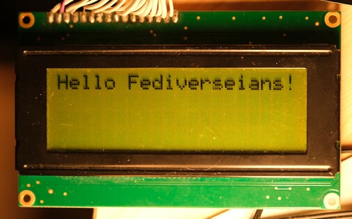 Photograph of display showing text. It reads "Hello Fediversians!". The display is old-fashioned, with each letter being in a cell of 5x8 dark pixels against a transflective green-ish background. The display is mounted in a dark brown bezel on a green printed copper board. At the top of the board, 16 wires are soldered which go out of frame. Each corner has a gold-plated screw hole.