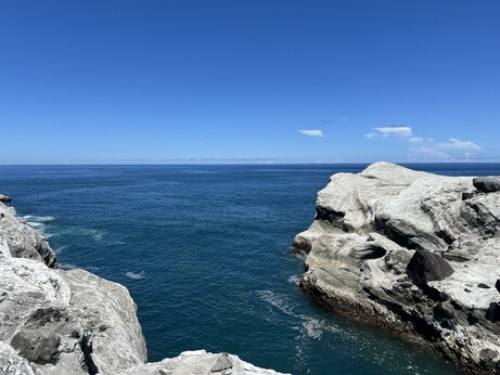 A vast ocean under a blue sky, with rugged rocky cliffs poking out into the water.