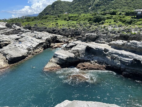 Aquamarine tide pools filling up with the surf among jagged rocks and a verdant hillside in the background.