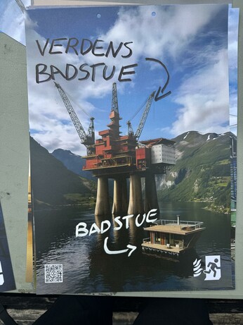 A view of a fiord with a floating sauna and an oil platform photoshopped hjust behind it.
The writing "badstue" with an arrow pointing to the sauna has been written with a white marker, while the writing "verdens badstue" and an arrow pointing to the platform has been written with a black marker.