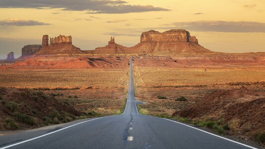 The view from the top of a small hill with a long straight road leading to the iconic red rock formations of Monument Valley under a vast western sky with a few layers of clouds.