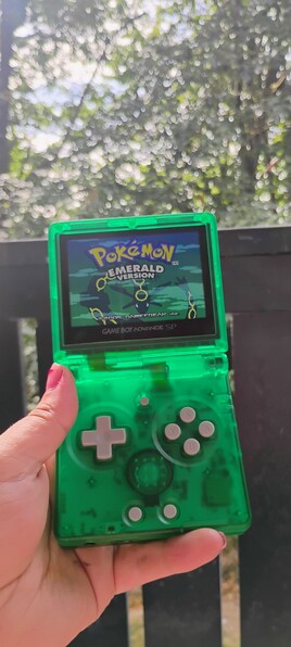 pokemon emerald title screen on my vibrant green translucent anbernic RG35XXSP. it's a gameboy sp inspired clamshell retro gaming device. leaves and trees in the background