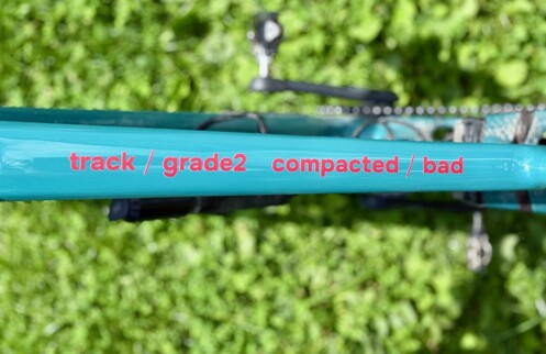 The image shows a close-up of a bicycle frame, specifically focusing on the top tube. The frame is painted in a bright turquoise color. On the frame, there is text written in red that reads “track / grade2  compacted / bad.” The background is blurred, but it appears to be an outdoor setting with greenery, suggesting that the photo was taken in a natural environment, possibly a park or a garden.